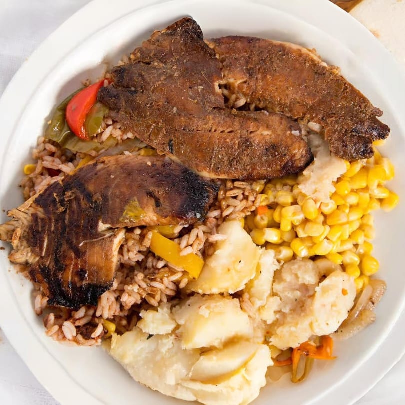 A Jamaican meal with brown stew fish, rice and peas, corn, and boiled dumplings on a white plate.