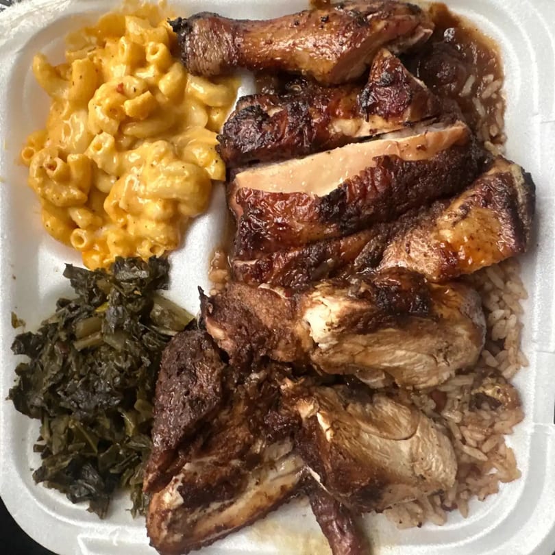 Jerk chicken with rice and peas, macaroni and cheese, and collard greens in a styrofoam container.