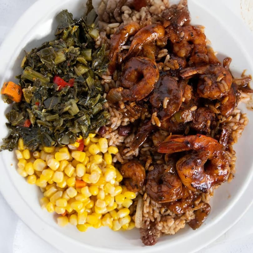 A Jamaican dish with brown stew shrimp, rice and peas, collard greens, and corn.